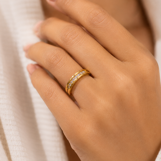 A close-up of a person's hand wearing a slim yellow gold ring with a row of small clear stones, set against a soft white textured garment.