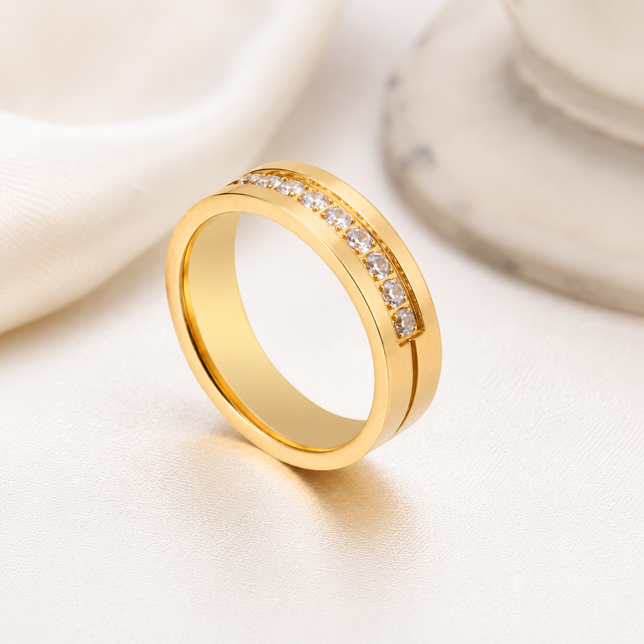 A gold band ring with a row of clear zircon stones along the top, displayed on a soft cream fabric background with a blurred marble object in the background.