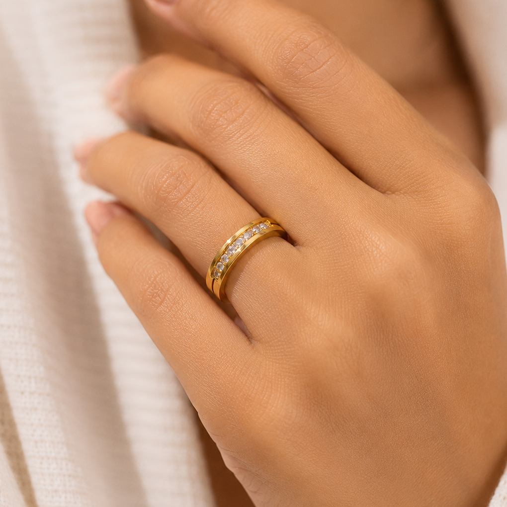 A close-up of a person's hand wearing a slim yellow gold ring with a row of small clear stones, set against a soft white textured garment.
