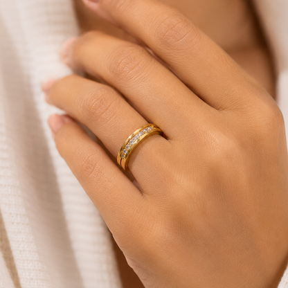 A close-up of a person's hand wearing a slim yellow gold ring with a row of small clear stones, set against a soft white textured garment.