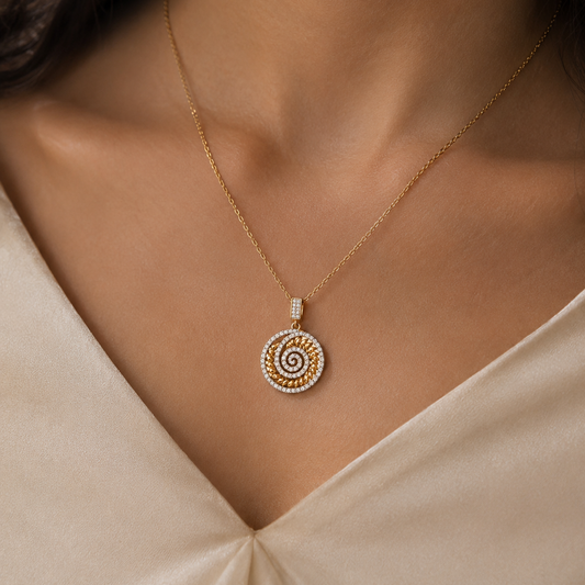 A close-up of a model wearing a delicate gold chain necklace with a circular spiral pendant set with small zircon stones, paired with a cream-colored satin top.