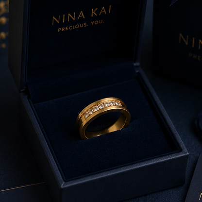 A gold ring with small clear zircon stones displayed in an open navy jewelry box, alongside matching navy gift boxes, cards, and ribbon tags branded NINA KAI on a dark styled background.